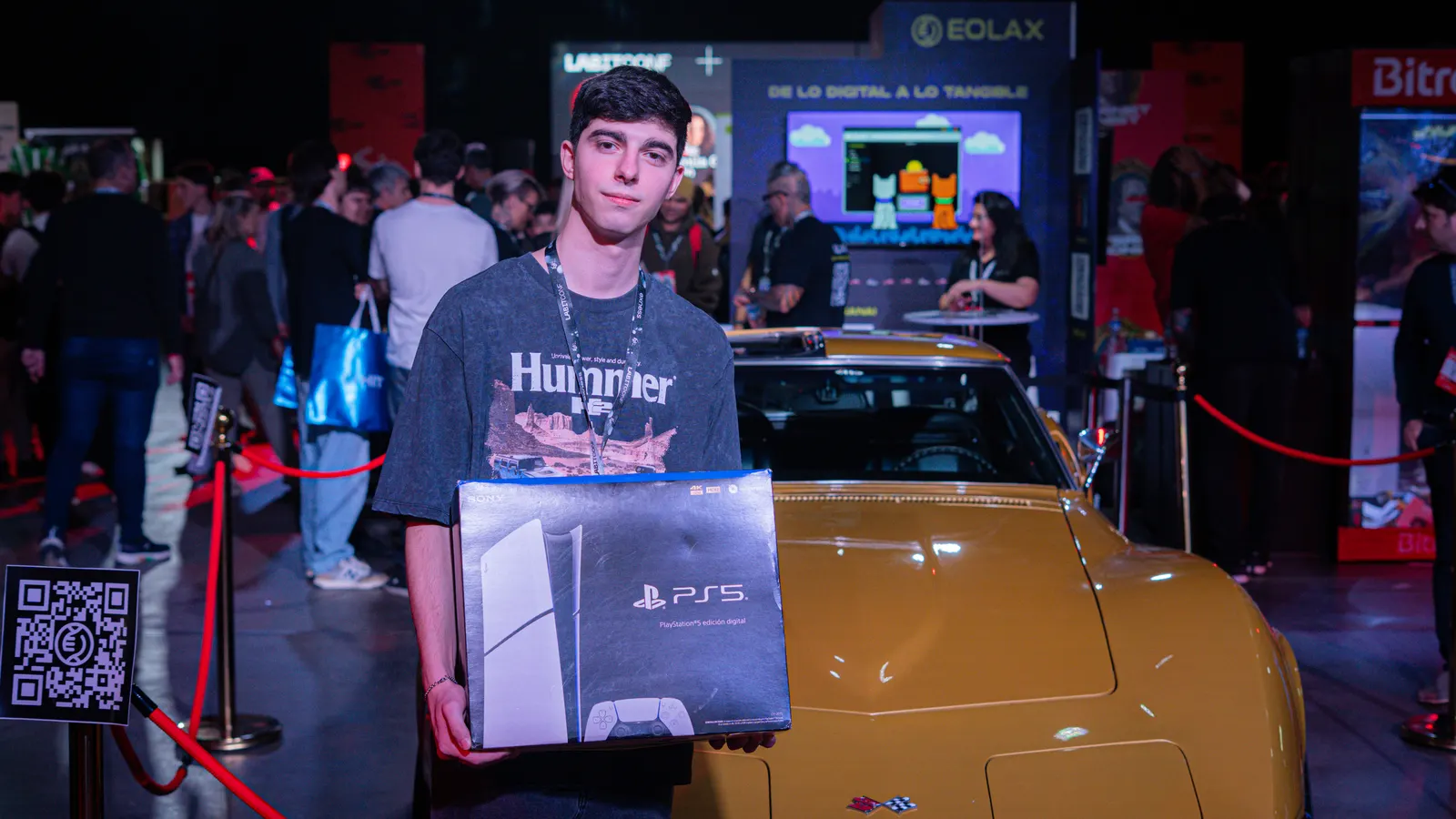 The winner holds the PlayStation 5 in front of the gold Corvette Stingray on display, the conference floor behind him.