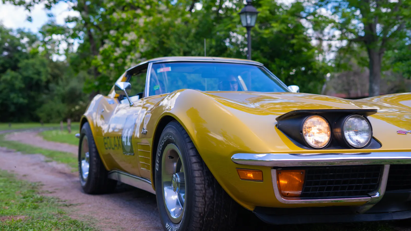 The Corvette Stingray seen head-on at three quarters, headlights lit, stopped on the dirt road.