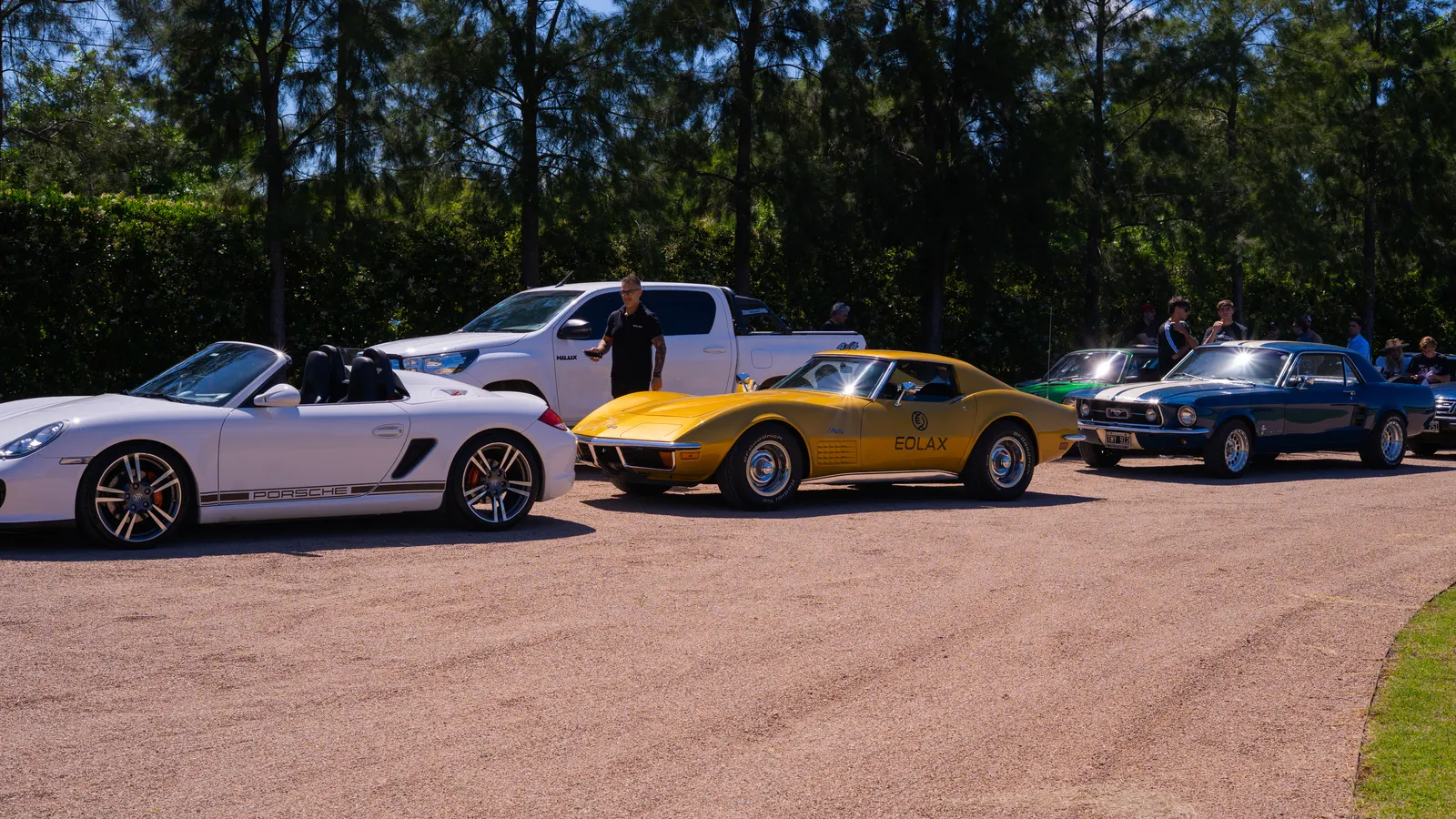 Classic cars parked on the gravel at the start: a white convertible, the gold Corvette and a blue coupé.