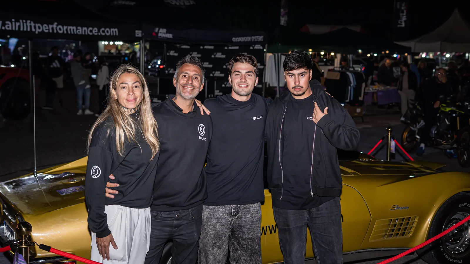 A group poses beside the gold Corvette on the grounds, after dark.