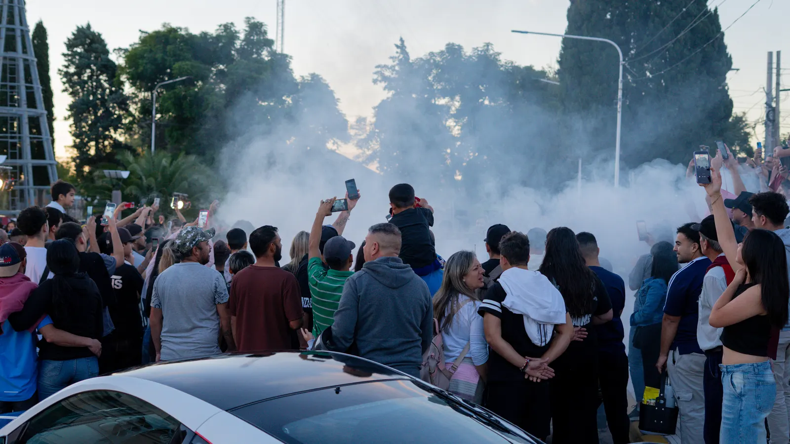 The crowd films with phones raised as a cloud of tyre smoke covers the track.