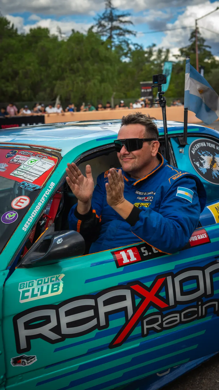 A driver waves from the window of his turquoise drift car, ready to head onto the track.
