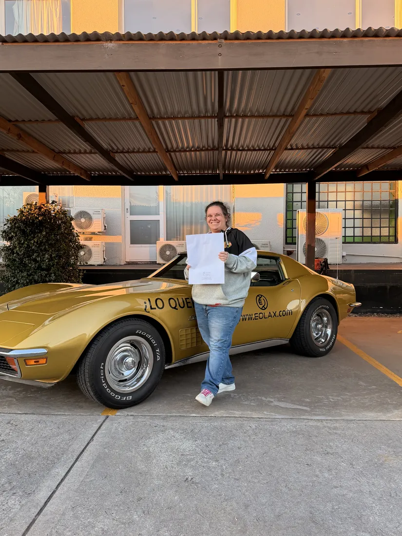 Laura P. sonríe sosteniendo el certificado frente al Corvette Stingray C3 dorado.